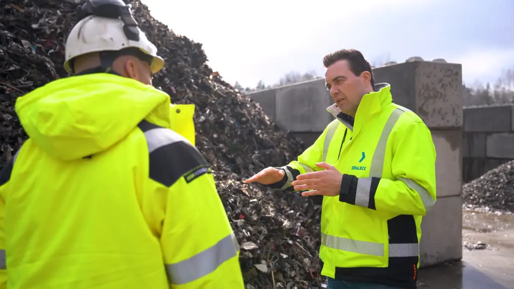 Twee mannen in gele jassen staan voor een hoop schroot te discussiëren.