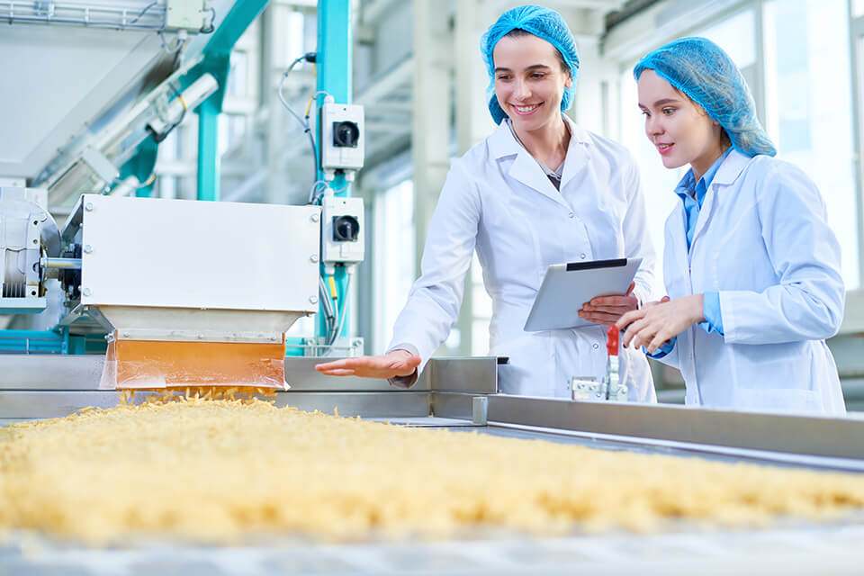 Twee vrouwen in laboratoriumjassen inspecteren een productielijn met bulkgoederen.