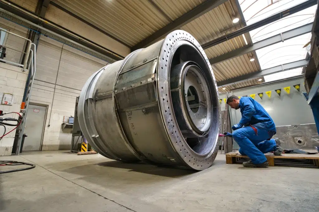 SPALECK-constructiemechanicus zit op zijn knieën bij een groot lasconstructieonderdeel en monteert onderdelen. Het onderdeel maakt deel uit van een groot gelast samenstel voor een gasturbine. De foto toont de productie bij SPALECK.
