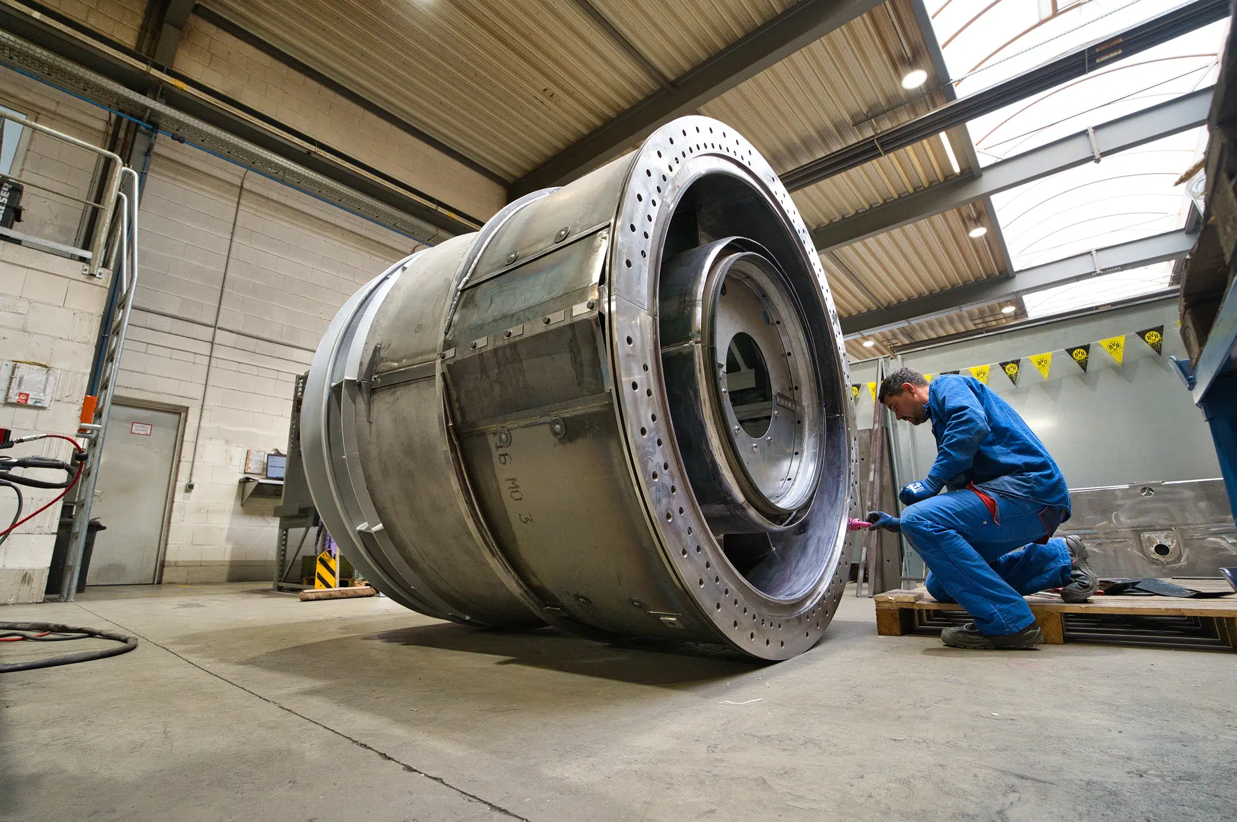 SPALECK-constructiemechanicus zit op zijn knieën bij een groot lasconstructieonderdeel en monteert onderdelen. Het onderdeel maakt deel uit van een groot gelast samenstel voor een gasturbine. De foto toont de productie bij SPALECK.