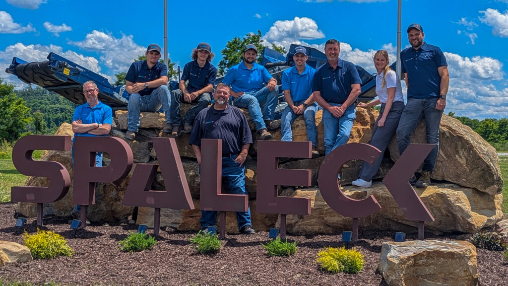 Groepsfoto van Spaleck USA bij het Spaleck-logo in Charleroi ter gelegenheid van de opening van de vestiging, met een blauwe lucht op de achtergrond